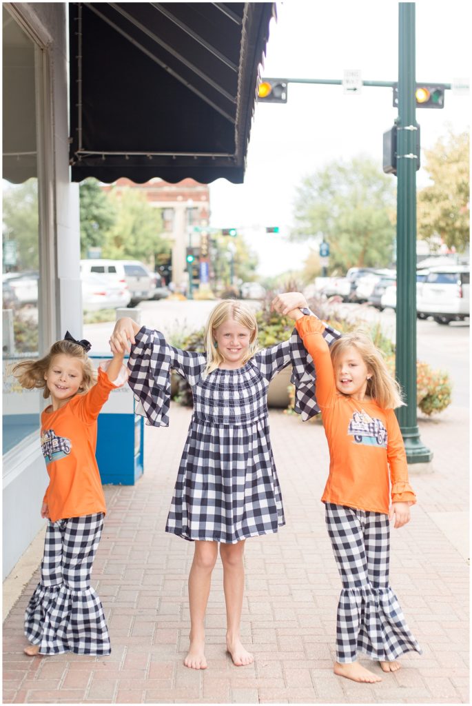 Sweet sisters are hand in hand walking on the sidewalk. 2 sisters are wearing matching orange shirts with a black and white checkered truck and matching black and white checkered flared pants. Older sister is wearing a long bell sleeve knee-length black and white checkered dress.