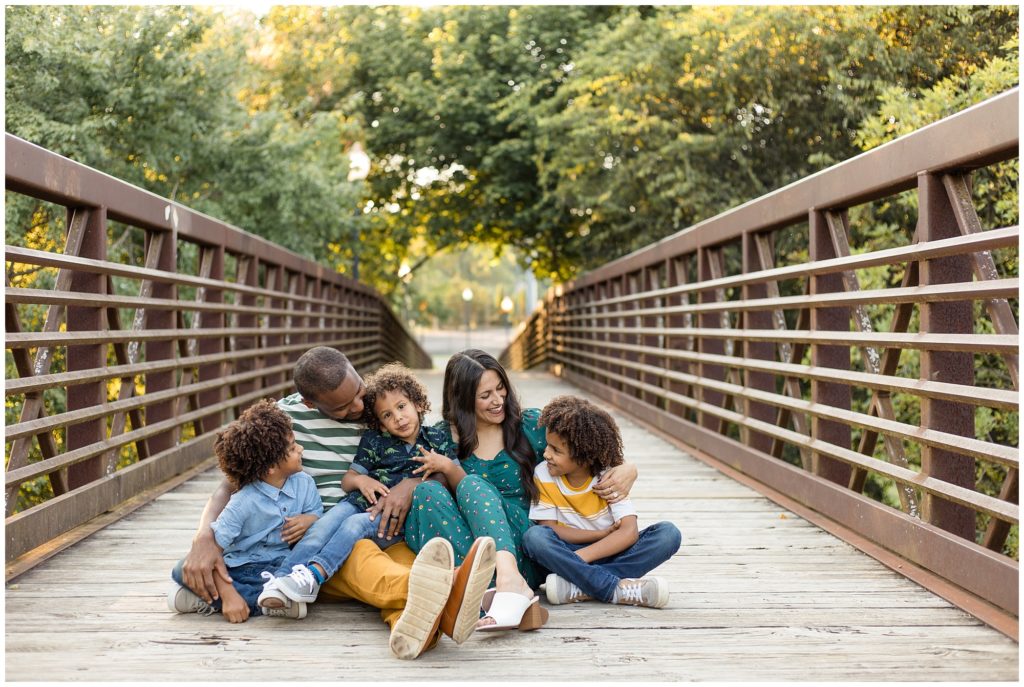 A beautiful family of 5 is seated on a rustic bridge. 1st brother is wearing a light blue polo and blue jeans. Dad is wearing a short sleeve green and white striped shirt and mustard yellow pants. 2nd brother is wearing a dark green shirt with light green dinosaurs and blue jeans. Mom is wearing a green jumper with a sweetheart neckline and floral print and white mule shoes. 3rd brother is wearing a mustard yellow and white striped short sleeve shirt and blue jeans. 