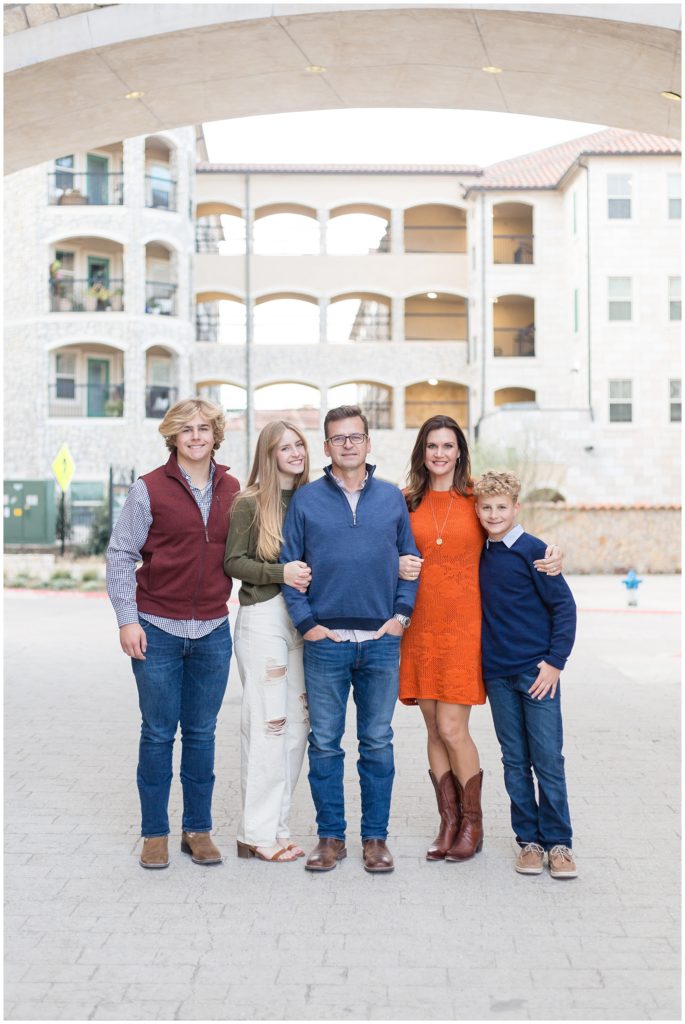 This gorgeous family of 5 is pictured standing in the center of this beautiful open structure with amazing archways in the background. 1st son is wearing a blue and white long sleeve button-up shirt with a maroon zip-up vest, blue jeans, and boots. Sister is wearing an olive green long sleeve shirt with white pants and brown sandals. Dad is wearing a long sleeve blue pullover shirt, blue jeans, and brown boots. Mom is wearing an orange knee-length dress with a slightly lighter orange floral detail and brown cowboy boots. 2nd son is wearing a long sleeve blue sweater with a white collared shirt popping out of the top, blue jeans, and tan shoes. 