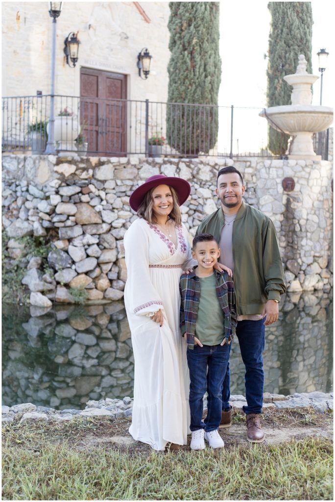 This gorgeous family of 3 is standing just in front of a stone wall and lake. Mom is wearing a long flowy dress with burgundy trim at the neckline, waist, and wrist. She is also wearing a matching burgundy hat. Son is wearing a sage shirt with army green and blue plaid shirt, blue jeans, and white sneakers. Dad is wearing a light color shirt with a sage green jacket, blue jeans, and brown boots. 