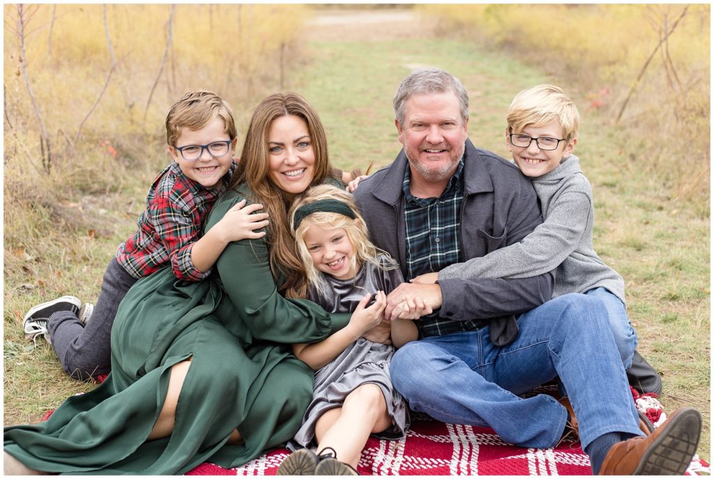 Beautiful family of 5 seated in an open field during this fall mini session. 1st son is wearing a red, green, and gray plaid shirt, grey pants, and black converse. Mom is wearing a gorgeous long-sleeve satin dress. The daughter is wearing short sleeve grey dress. Dad is dressed in a green, blue, and white plaid shirt with a grey jacket, blue jeans, and brown shoes. 2nd son is wearing a grey pullover and blue jeans.