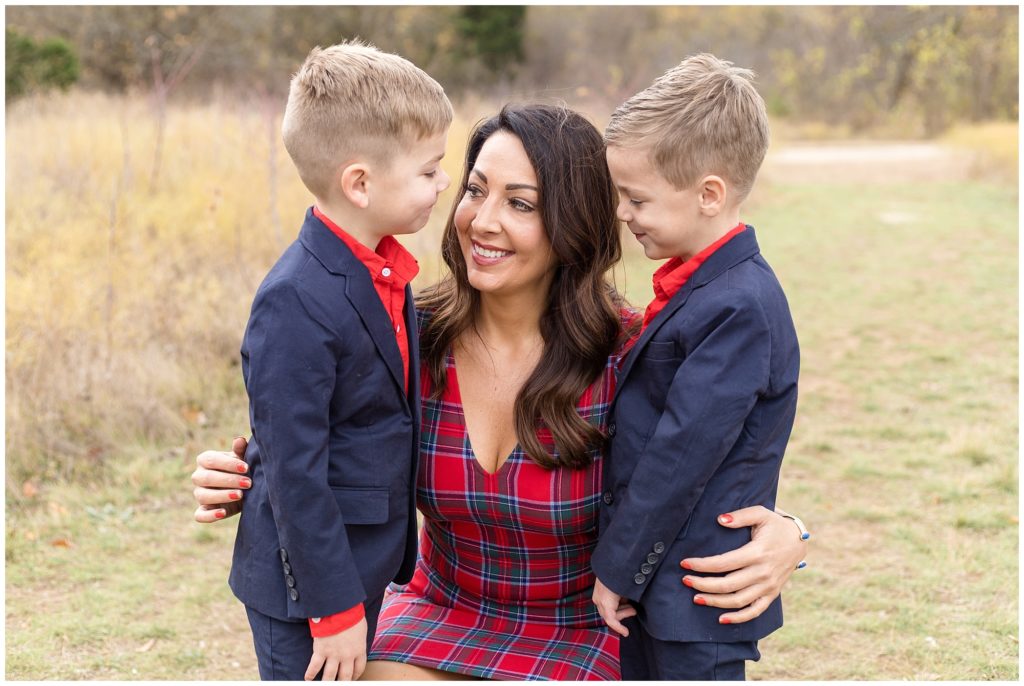 Mom and her 2 sons are pictured in a beautiful open field. The boys are wearing matching navy suits with a red button-up shirt. Mom is wearing a red, green, white, and navy plaid dress.