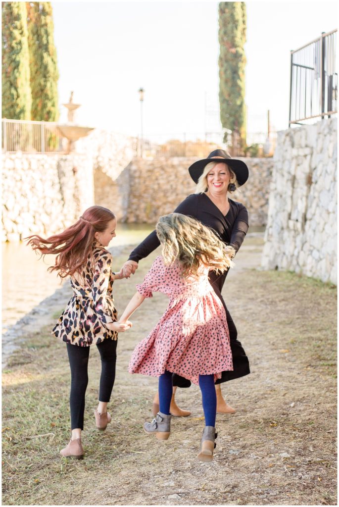 Mom and her daughters are dancing with beautiful stone walls in the background. Mom is wearing a long-sleeve full-length black dress with a wide brim hat and tan booties. The first daughter is wearing a long sleeve leopard print blouse, and black leggings taupe booties. The second daughter is wearing a short sleeve pink dress with black print, blue leggings, and grey booties.