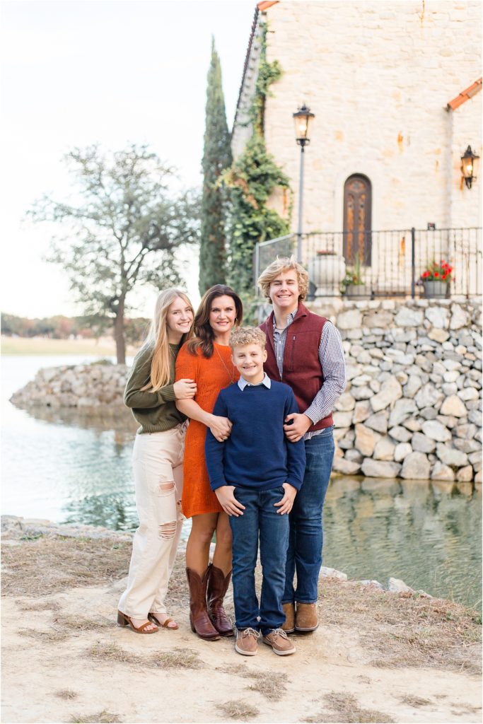 Mom and her 3 kids are standing at the edge of a pond with a gorgeous stone wall and chapel at Adriatica. Daughter is wearing a long sleeve olive green shirt and white jeans. Mom is waering a short sleeve knee length dress and cowboy boots. 1st son is wearing a long sleeve navy blue sweater with a collar underneath and blue jeans. 2nd son is wearing a long sleeve button up checkered shirt and  a rust colored vest.