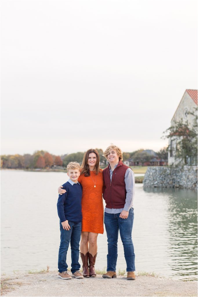 Mom and her sons are standing just in front of a pond with a gorgeous stone wall and chapel in the background. Mom is wearing a short sleeve knee length dress and cowboy boots. 1st son is wearing a long sleeve navy blue sweater with a collar underneath and blue jeans. 2nd son is wearing a long sleeve button up checkered shirt and  a rust colored vest.