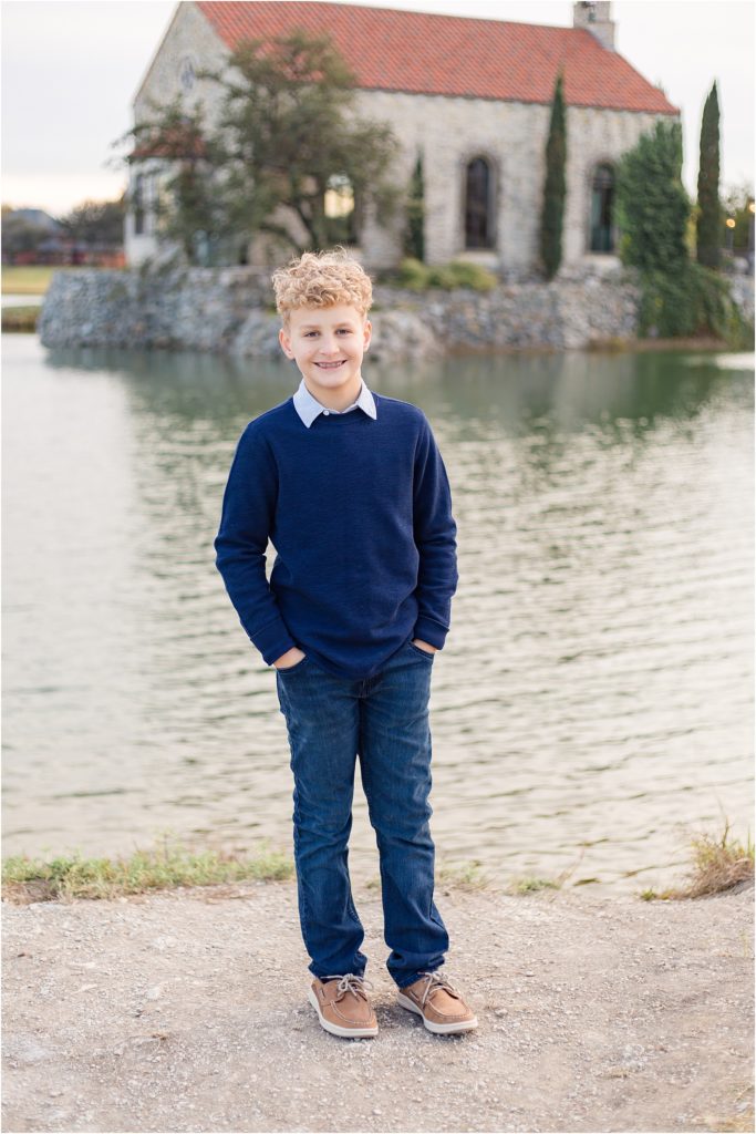 Son is standing on the edge of a gorgeous pond with a beautiful stone wall and chapel. He is wearing a long sleeve navy blue sweater with a collar underneath and blue jeans. 