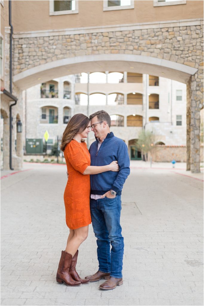Mom and dad are standing head to head hugging with a gorgeous stone archway in the back ground. Mom is wearing a short sleeve knee length dress and cowboy boots. Dad is wearing a long sleeve navy pull over sweater and blue jeans