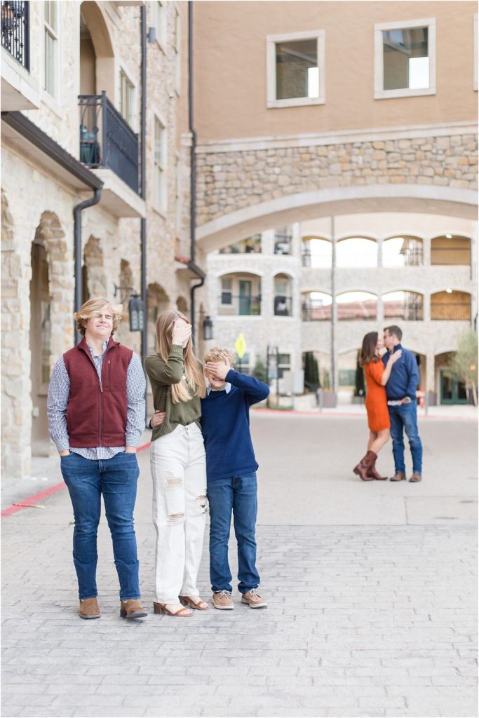 Family of 5 are standing on a stone street with a gorgeous stone archway in the background with gorgeous stone wall on the side. Mom is waering a short sleeve knee length dress and cowboy boots. 1st son is wearing a long sleeve navy blue sweater with a collar underneath and blue jeans. 2nd son is wearing a long sleeve button up checkered shirt and  a rust colored vest. Dad is wearing a long sleeve navy pull over sweater and blue jeans