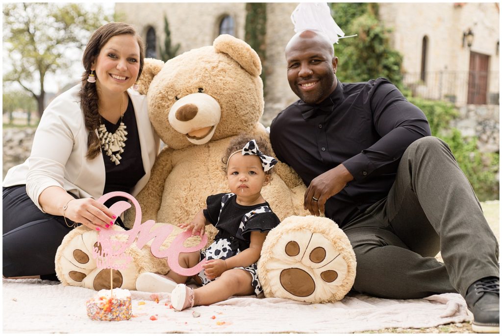 Sweet family of 3 are seated on a white blanket with giant teddy bear. Mom is wearing black pants and a black shirt with a white jacket. She is wearing a large white necklace and matching dangle earrings. Dad is wearing a long sleeve button up shirt wand grey pants. Daughter is wearing a a black shirt with ruffle sleeves and a black and white cow print jumper with a matching bow.
Adriatica Village | McKinney, TX