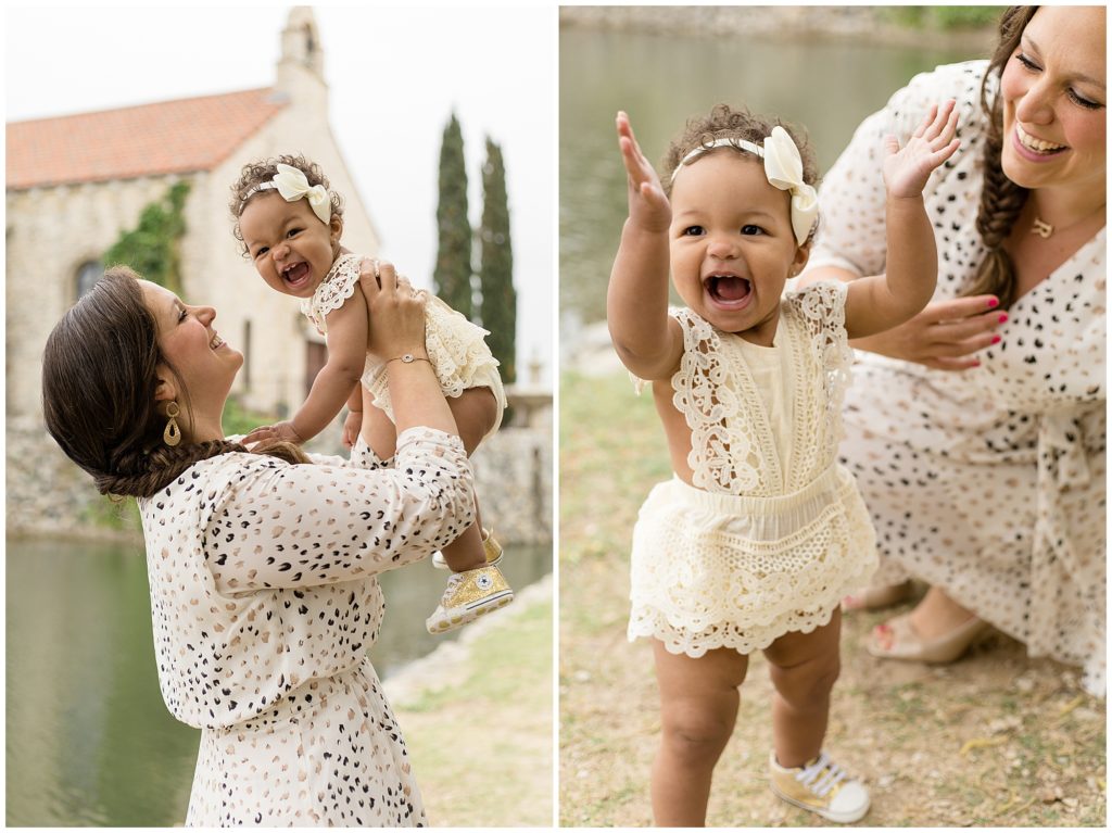 Mom and daughter are standing just in front of a stone building, stone wall and small pond. Mom is wearing a white dress with black and gold print. Daughter is wearing a cream colored sleeveless lace trim romper with a a matching bow.