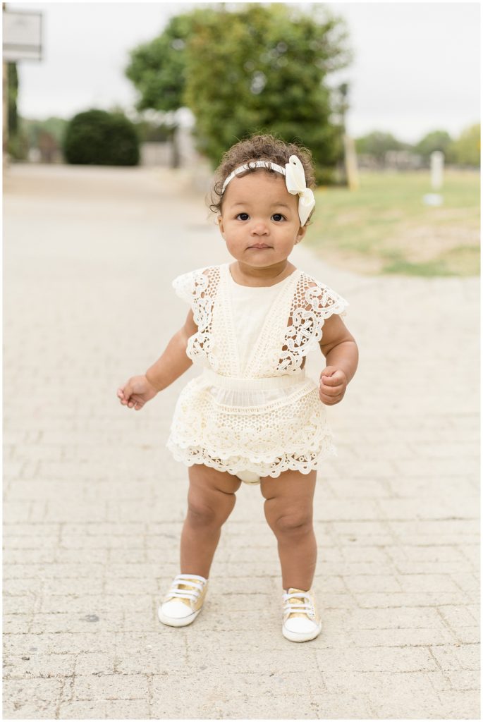 Adorable baby girl is wearing a sleeveless cream colored lace trim romper and matching bow. She is pictured on a paved path with a large tree in the background.