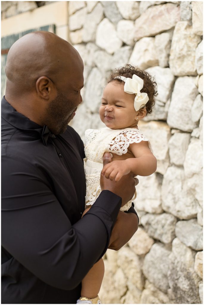 Dad and daughter are standing near a stone wall. Dad is wearing a long sleeve button up black shirt. Baby girl is wearing a sleeveless cream colored lace trim romper and matching bow. 
Adriatica Village | McKinney, TX
