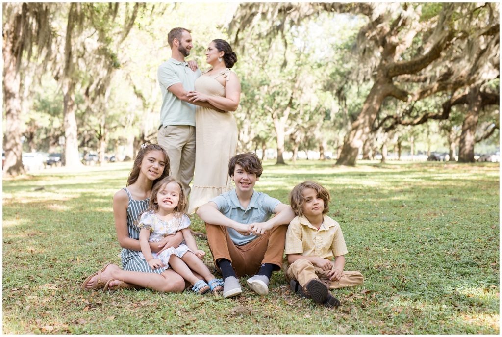Family of 6 surrounded by gorgeous trees. The 4 kids are seated. 1st daughter is wearing a sleeveless blue and white striped dress and tan sandals. 2nd daughter is wearing a white dress with a floral print and blue sandals. 1st son is wearing a light blue short sleeve polo and brown pants. 2nd son is wearing a yellow and white checkered shirt and brown pants. Dad is wearing a light green short sleeve polo and khaki pants. Mom is wearing a cream-colored sleeveless dress with straps tied at the shoulders | Daffin Park Family Session