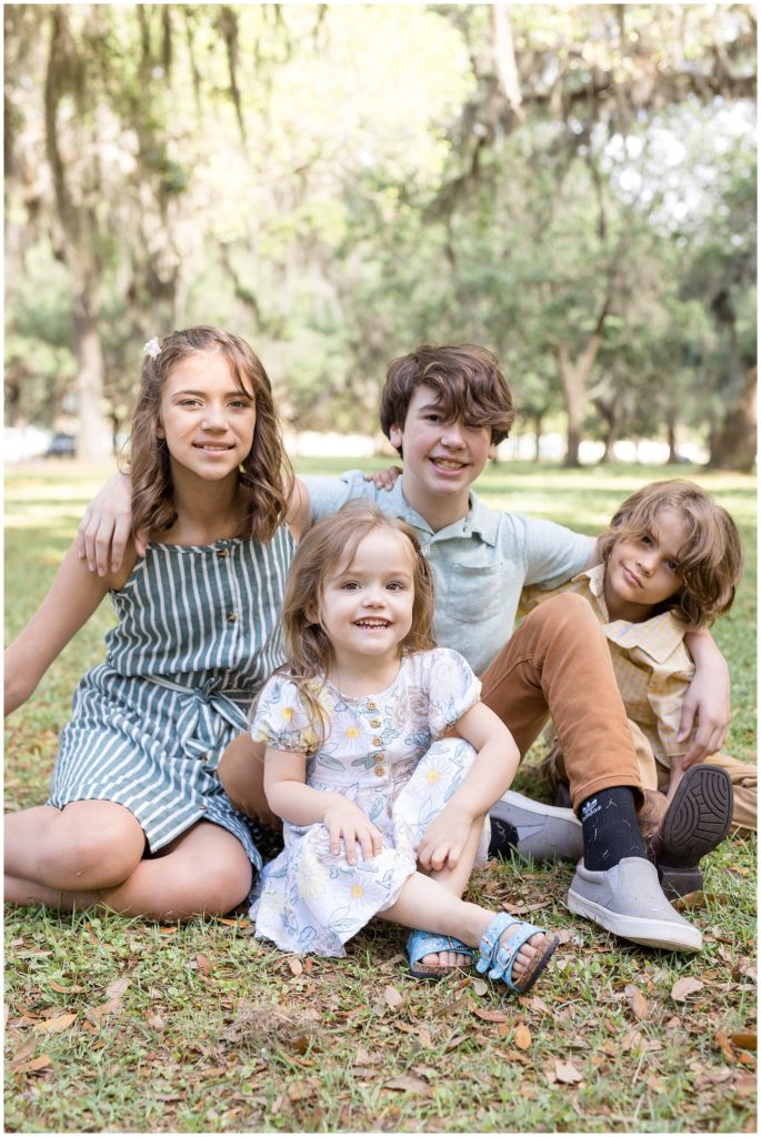 Kids are sitting with arms around each other and beautiful trees are just in the background.1st daughter is wearing a sleeveless blue and white striped dress and tan sandals. 2nd daughter is wearing a white dress with a floral print and blue sandals. 1st son is wearing a light blue short sleeve polo and brown pants. 2nd son is wearing a yellow and white checkered shirt and brown pants | Daffin Park Family Session