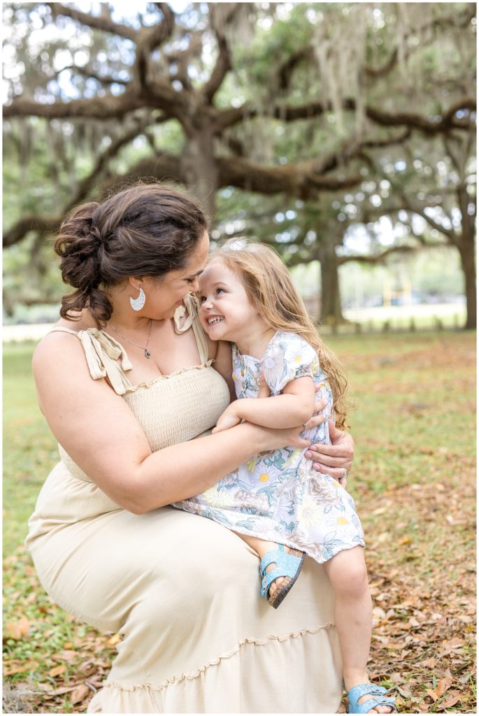 Mom is holding her adorable daughter while they both look at each other. Mom is wearing a cream-colored sleeveless dress that ties at the shoulders. The daughter is wearing a white dress with a large floral print and blue sandals. 
