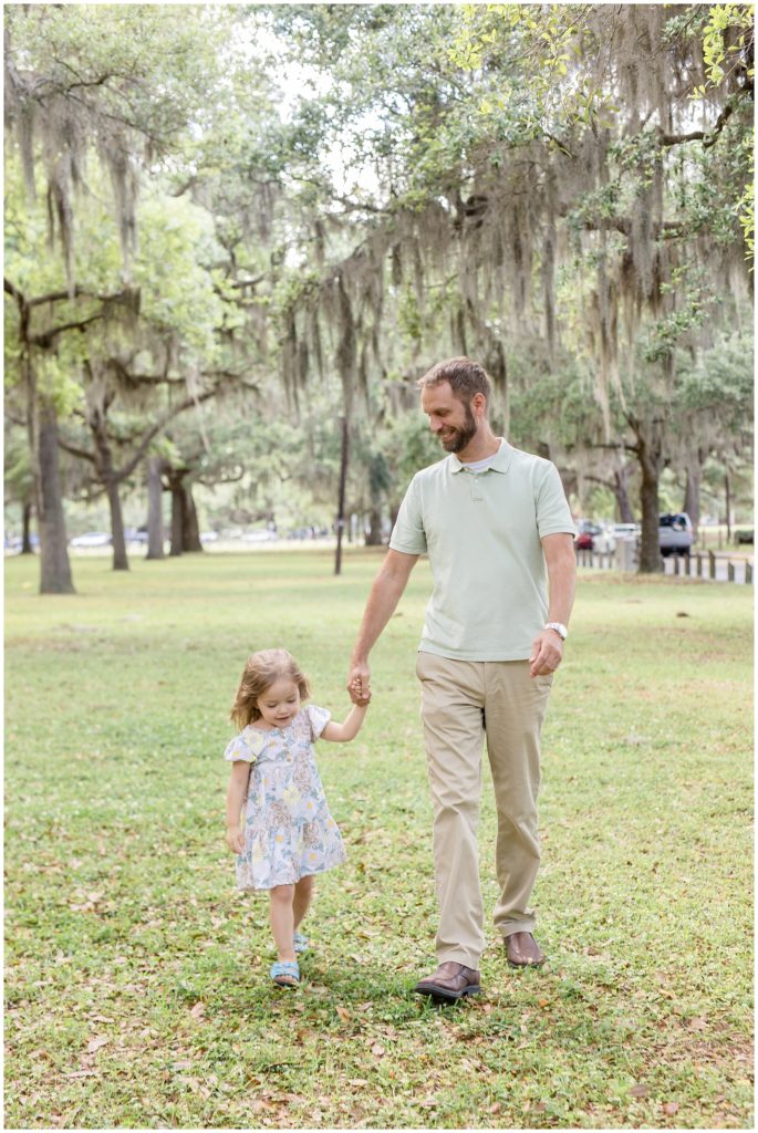 Dad and daughter are walking hand in hand between a gorgeous line of hanging trees. The daughter is wearing an adorable white dress with a large floral print and blue sandals. Dad is wearing a light green short sleeve polo with khaki pants and brown shoes.