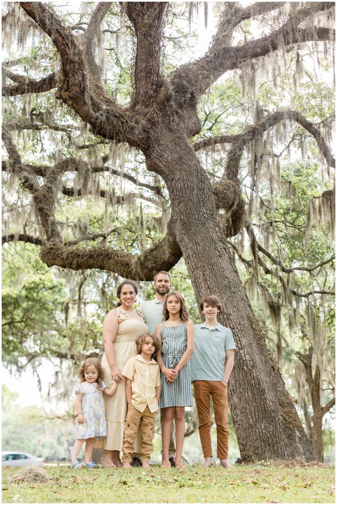Amazing shot of this beautiful family of 6 standing in front of a huge hanging trees. 1st daughter is wearing a sleeveless blue and white striped dress and tan sandals. 2nd daughter is wearing a white dress with a floral print and blue sandals. 1st son is wearing a light blue short sleeve polo and brown pants. 2nd son is wearing a yellow and white checkered shirt and brown pants. Dad is wearing a light green short sleeve polo and khaki pants. Mom is wearing a cream-colored sleeveless dress with straps tied at the shoulders | Daffin Park Family Session