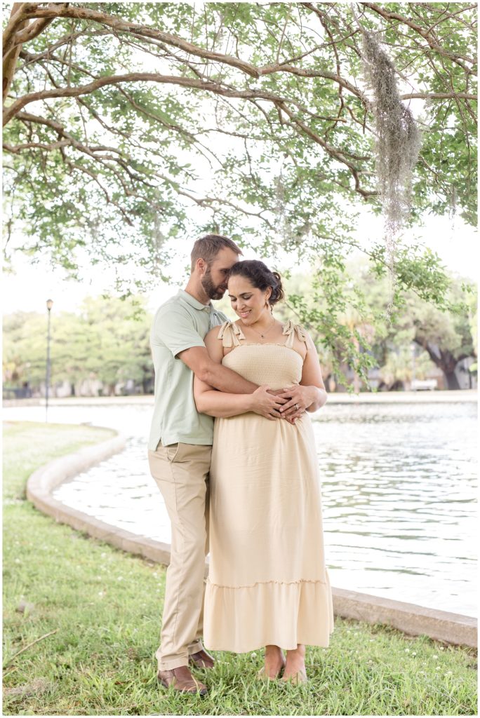 Mom and dad are sharing a hug while standing with a lake and beautiful hanging trees. Dad is wearing a light green short sleeve polo and khaki pants. Mom is wearing a cream-colored ankle-length dress that ties at the shoulders | Daffin Park Family Session