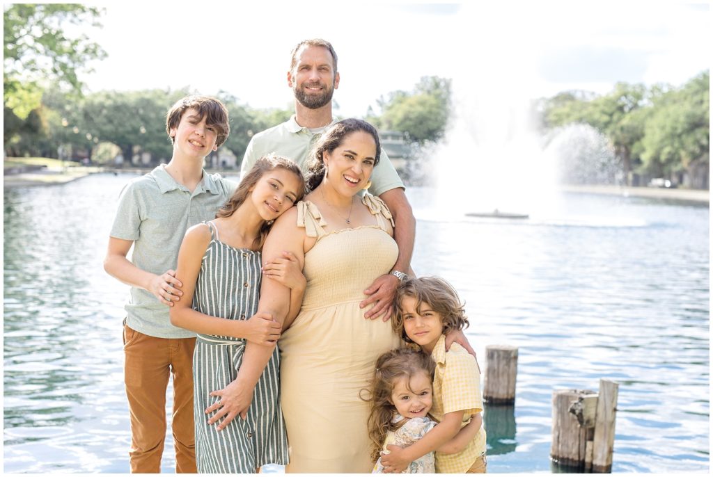 A gorgeous family of 6 is standing in front of the lake and fountain. 1st daughter is wearing a sleeveless blue and white striped dress and tan sandals. 2nd daughter is wearing a white dress with a floral print and blue sandals. 1st son is wearing a light blue short sleeve polo and brown pants. 2nd son is wearing a yellow and white checkered shirt and brown pants. Dad is wearing a light green short sleeve polo and khaki pants. Mom is wearing a cream-colored sleeveless dress with straps tied at the shoulders.
Daffin Park Family Session