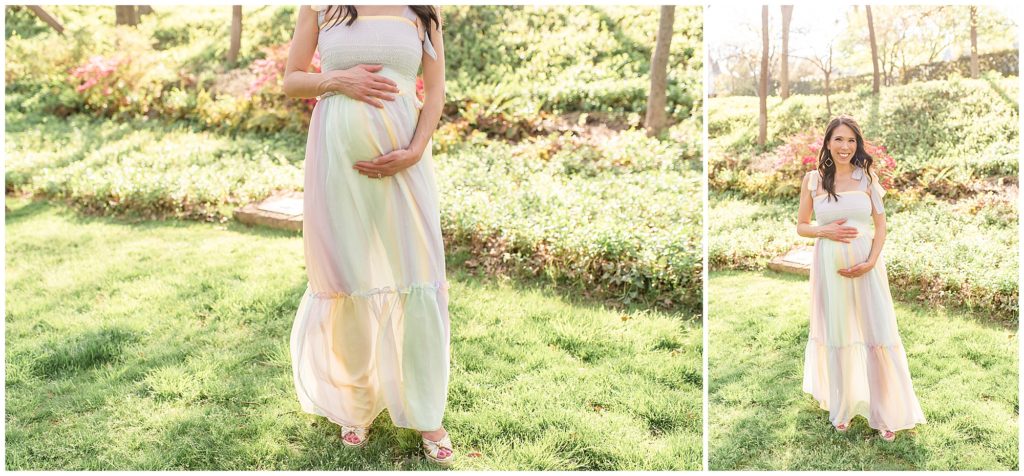 Expectant mom is standing in a beautiful grassy field holding her belly. Mom is wearing a gorgeous long sleeveless multi colored light pink, blue and yellow flowing dress tied at the shoulders.