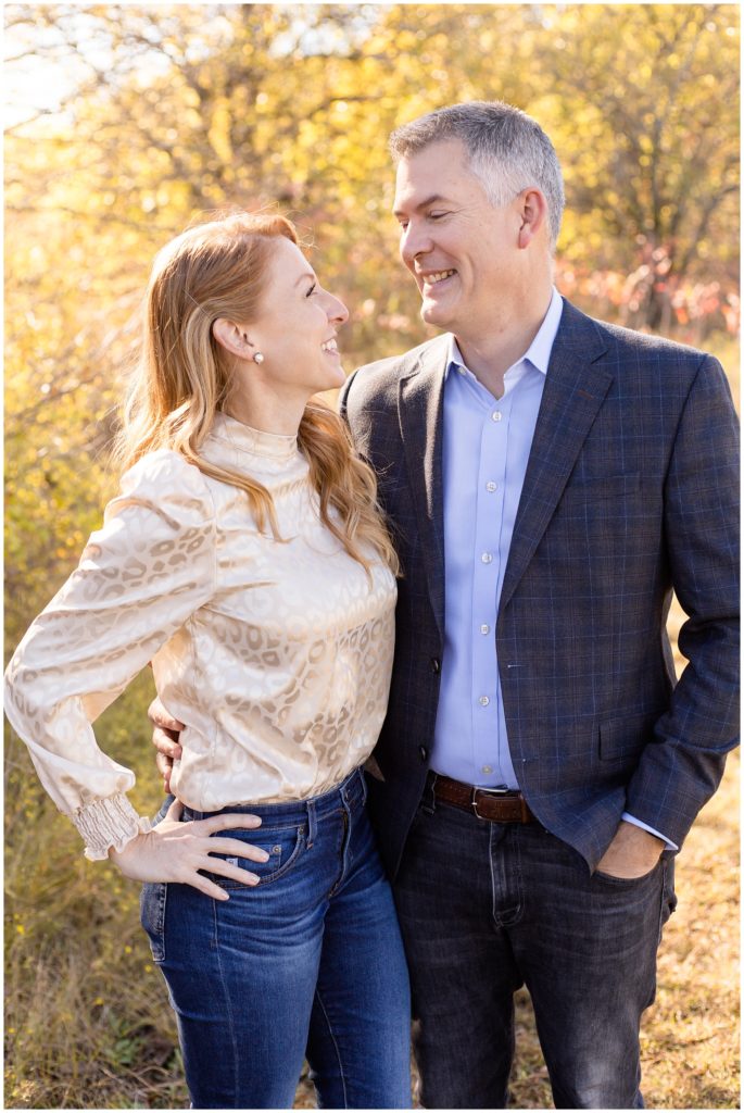 Mom and dad are standing surrounded by beautiful fall trees. Mom is wearing a blush pink metallic animal print long sleeve shirt and blue jeans. Dad is wearing a grey blazer with blue stripes and a light blue button-up shirt.
Family Portrait Session | Wisp + Willow Photography Co.