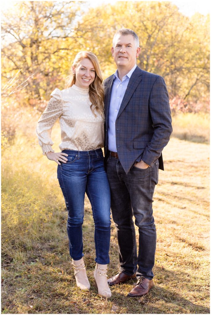 Mom and dad are standing surrounded by beautiful fall trees. Mom is wearing a blush pink metallic animal print long sleeve shirt and blue jeans. Dad is wearing a grey blazer with blue stripes and a light blue button-up shirt.
Family Portrait Session | Wisp + Willow Photography Co.