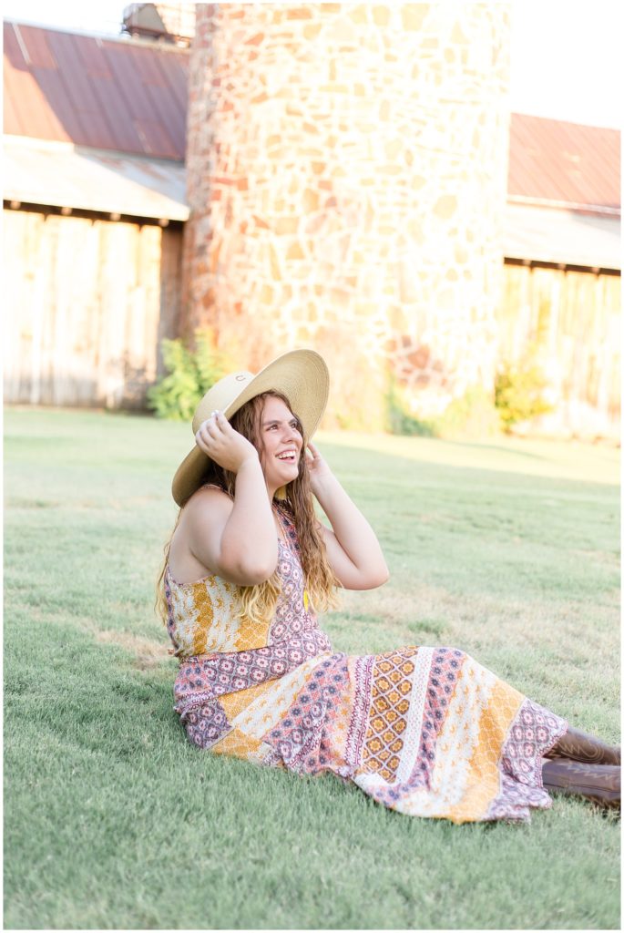 The client is seated in front of a rustic stone and wooden building. She is wearing a beautiful long, sleeveless multi-colored bohemian style dress with cowboy boots and a wide brim hat, and a long necklace with a bright yellow pendant and dangle fringe earrings.
