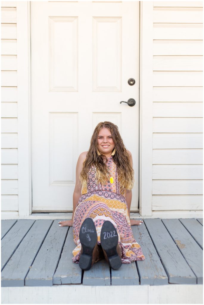 The client is seated on the porch of this white farmhouse. She is wearing a beautiful long, sleeveless multi-colored bohemian style dress with cowboy boots and a long necklace with a bright yellow pendant and dangle fringe earrings.