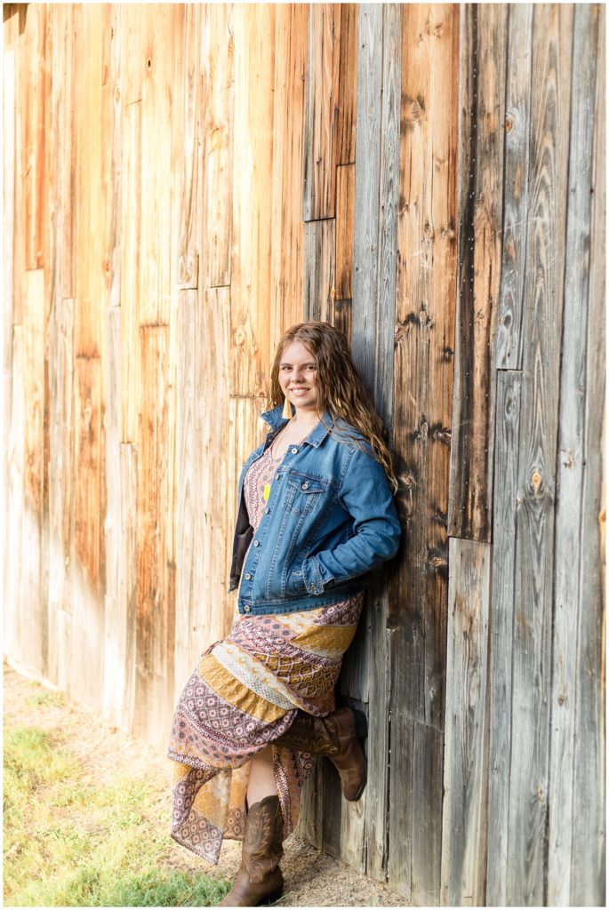 The client is leaning against a beautiful rustic wooden exterior wall in this Frisco Heritage Center Senior Session. She is wearing a beautiful long, sleeveless multi-colored bohemian style dress with cowboy boots, a jean jacket, and a long necklace with a bright yellow pendant and dangle fringe earrings.