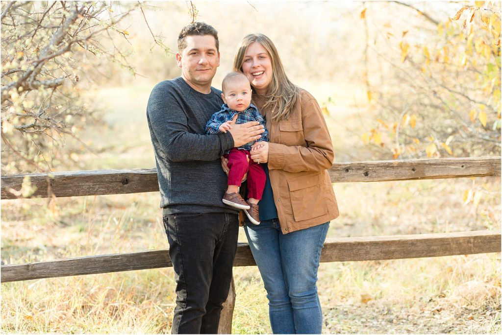 Precious family of 3 are standing at a rustic wooden fence and tress to each side. Dad is wearing a grey long sleeve sweater and black pants. Mom is wearing a brown jacket and blue jeans. Son is wearing a blue plaid long sleeve shirt and red pants.
