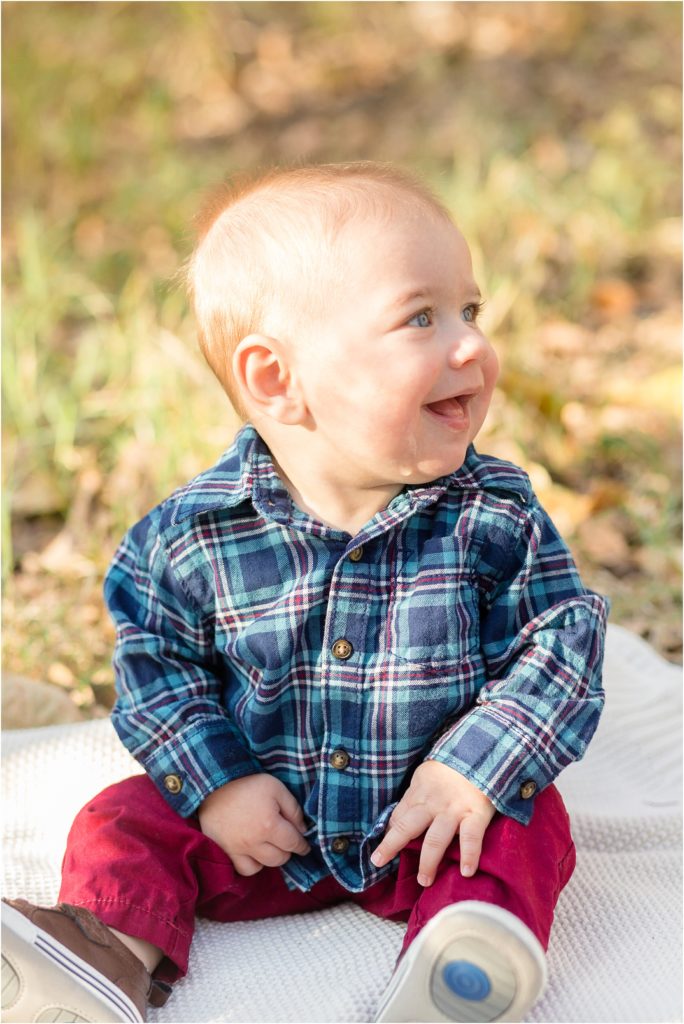 Sweet baby boy is seated on a white blanket at Arbor Hills. He is wearing a blue and red plaid long sleeve button up shirt and red pants.