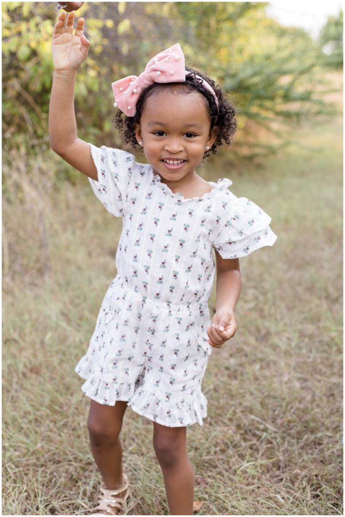 The precious little girl is walking through an open field with trees just in the background in Frisco Commons Location Spotlight session She is wearing a white flutter sleeve romper with a tiny purple flower print and a large pink bow with pearl accents and pearl earrings.