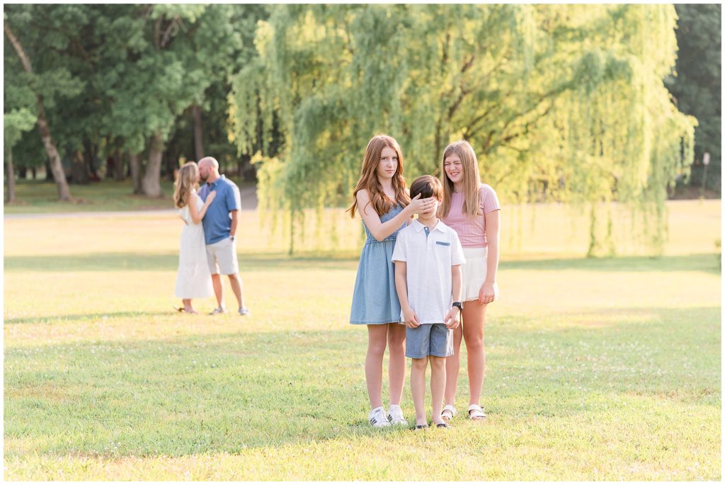 Family is standing in an opening with a beautiful hanging tree in the background. Daughter is wearing a blush pink short sleeve shirt with a white flutter skirt. Second daughtere is wearing a blue sleeveless knee length dress. Mom is wearing a sleeveless ankle length dress. Dad is wearing a short sleeve blue polo with khaki shorts. Son is dressed in a white short sleeve polo and blue shorts.