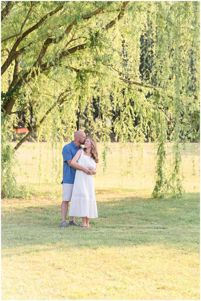 Mom and dad are standing just beneath a gorgeous hanging tree. Mom is wearing a sleeveless ankle length dress. Dad is wearing a short sleeve blue polo with khaki shorts. .