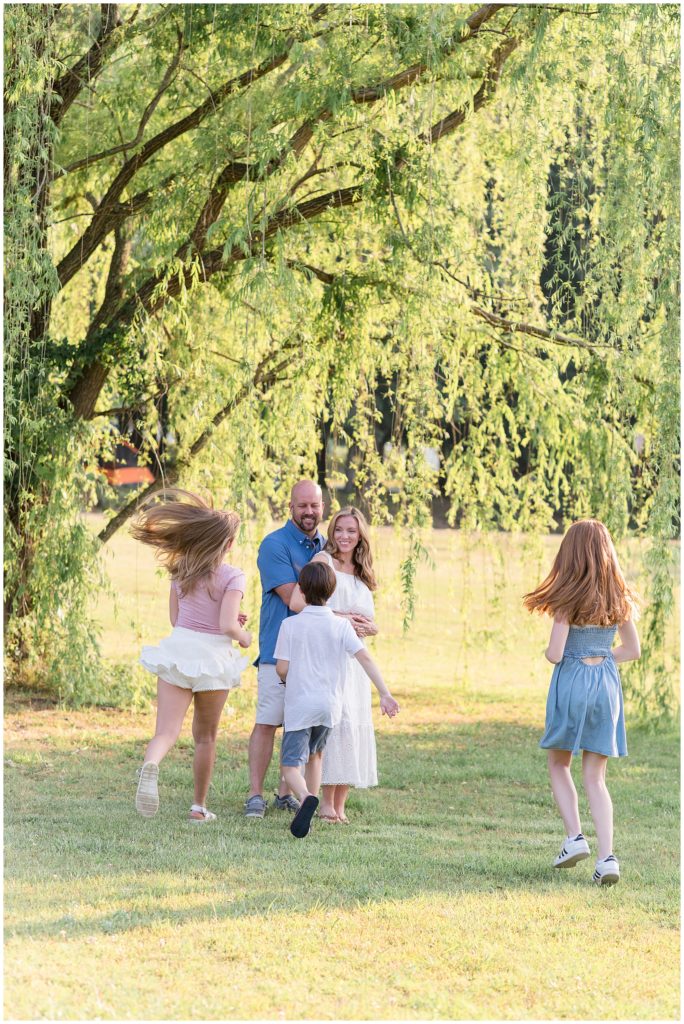 Family of 5 pictured with kids running to parents who are standing just beneath a hanging tree. Daughter is wearing a blush pink short sleeve shirt with a white flutter skirt. Second daughtere is wearing a blue sleeveless knee length dress. Mom is wearing a sleeveless ankle length dress. Dad is wearing a short sleeve blue polo with khaki shorts. Son is dressed in a white short sleeve polo and blue shorts.