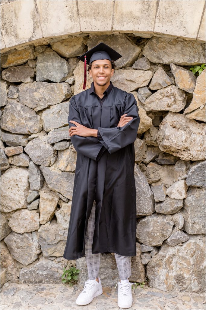 This high school senior is standing against an amazing stone wall. He is wearing his cap and gown for his senior graduation. 