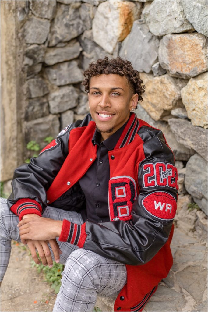 This high school senior is wearing his black and red letterman jacket. With a black shirt and black and grey plaid pants. He is seated in front of a large stone wall. 