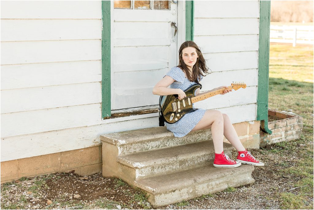 This high school senior chose to bring her guitar to her senior session. She is seated on stone steps in front of a rustic white house and green trim. 
