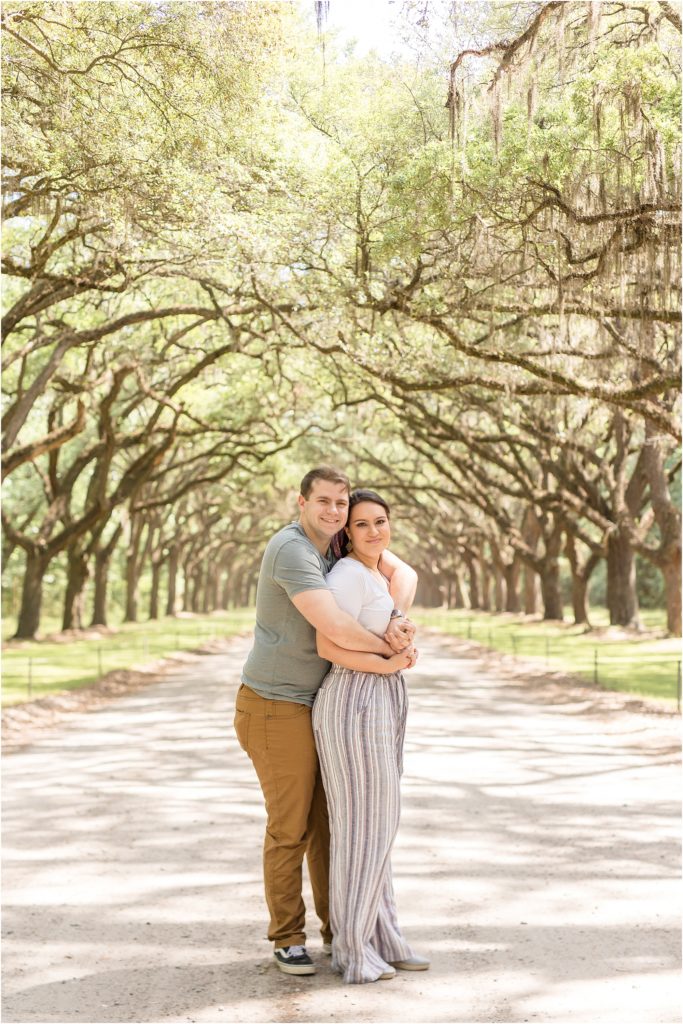 Husband and wife are standing on a path beneath an archway of trees. Husband is standing behind wife hugging her. Wife is wearing a short sleeve white shirt and a multi-colored striped pants. Husband is wearing a sage green short sleeve shirt and brown pants.