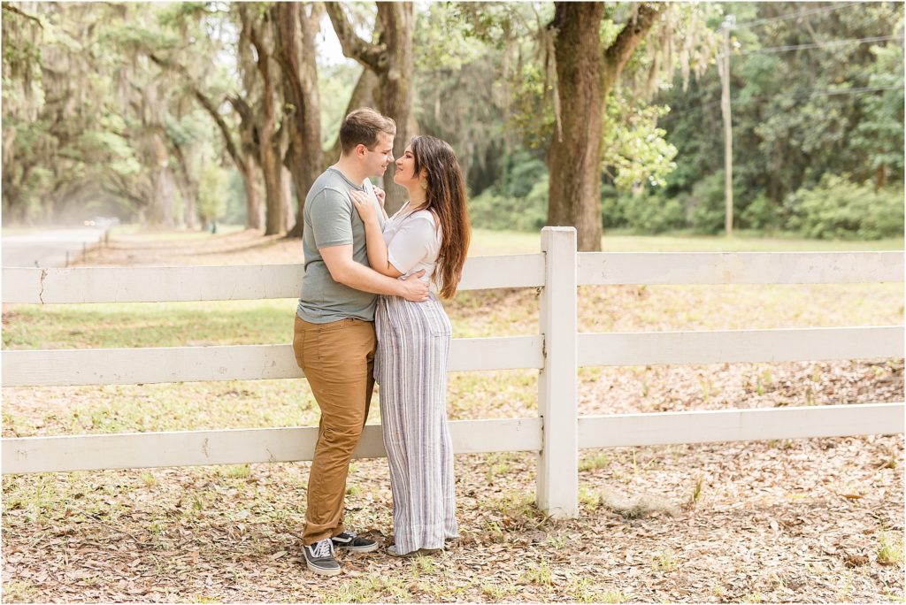 Husband and wife are standing in front of a white wooden fence and beautiful hanging trees. They are looking at each other hugging. Wife is wearing a short sleeve white shirt and a multi-colored striped pants. Husband is wearing a sage green short sleeve shirt and brown pants.