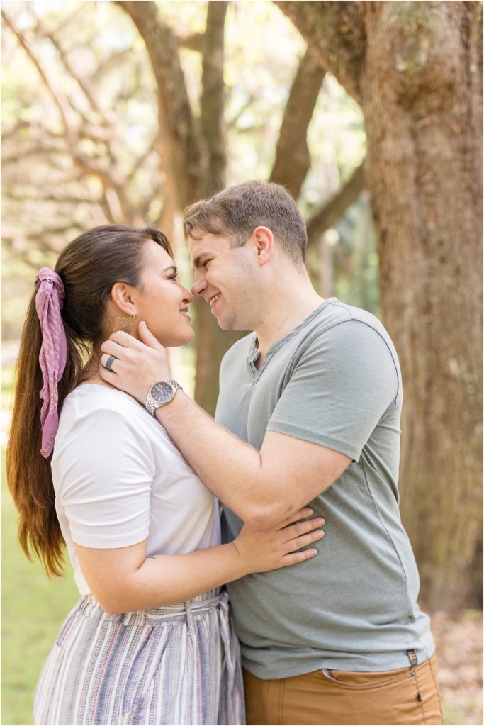 Husband and wife are standing nose to nose hugging with huge trees in the background. Wife is wearing a short sleeve white shirt and a multi-colored striped pants with her hair in a ponytail tied with a purple scarf.  Husband is wearing a sage green short sleeve shirt and brown pants.