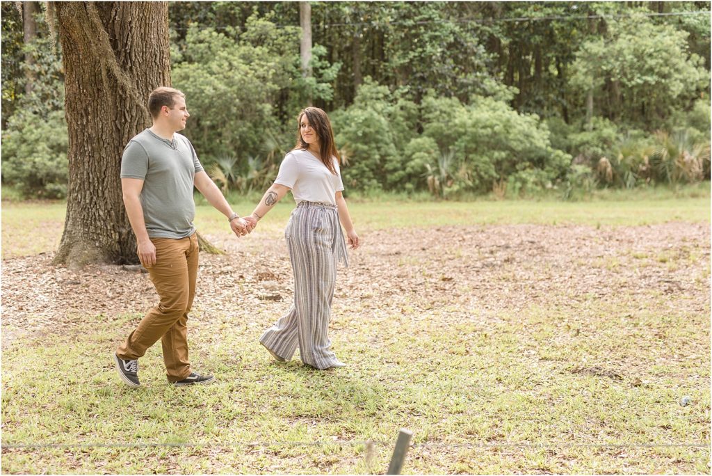 Husband and wife are holding hands, looking at each other walking trough a field with a huge tree and gorgeous tree-line in the background.  Wife is wearing a short sleeve white shirt and a multi-colored striped pants. Husband is wearing a sage green short sleeve shirt and brown pants.