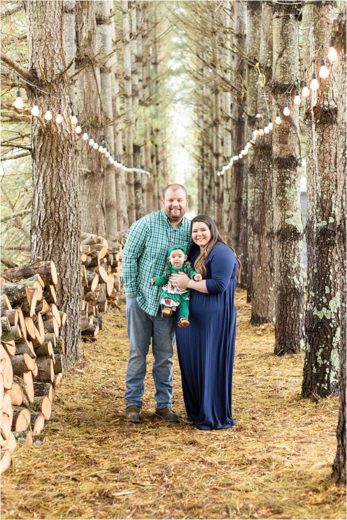 Family of 3 are standing in a tree farm. Dad is wearing a long sleeve button up green plaid shirt and blue jeans. Baby is waring a green long sleeve shirt with a ruffle at the bottom and red and white accents  and matching green pants. Mom is waring long sleeve ankle length navy dress. 