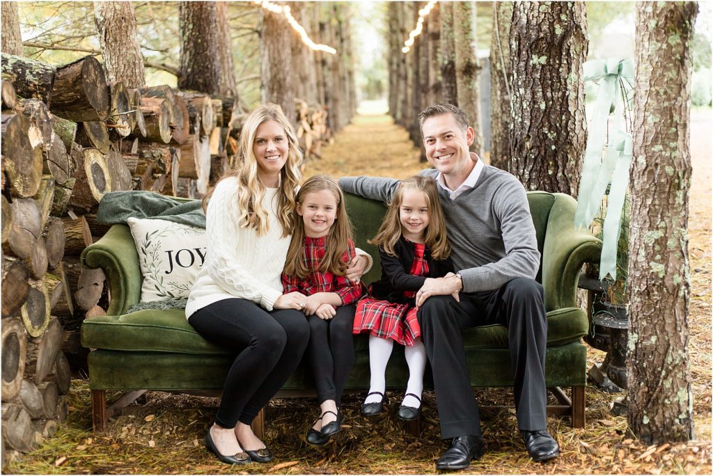 Gorgeous family of 4 is seated on a green velvet couch in a tree farm. Mom is waring a white sweater and black pants and black flats. 1st daughter is wearing a red and black paid tunic, black leggings and ballet flats. 2nd sister is wearing a red and black plaid dress with a black cardigan, white tights and black ballet flats. Dad is wearing a long sleeve grey sweater with a white collar peeking out of the collar of the grey sweater, black slacks and black shoes. 