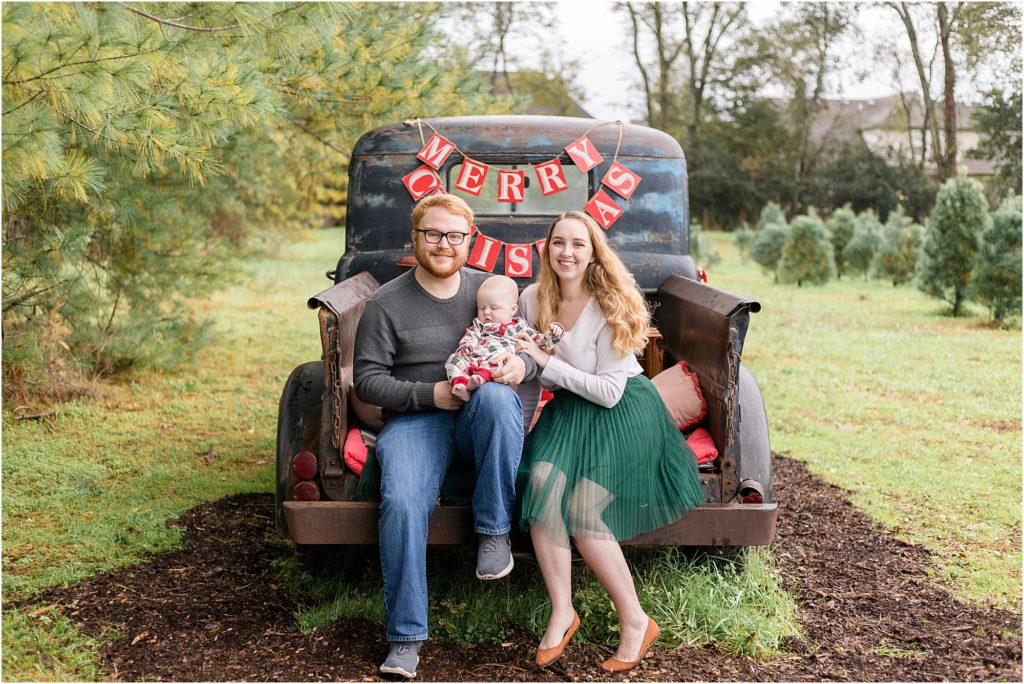 Precious family of 3 are seated on the back of a rustic vintage truck. Dad is wearing a long sleeve grey sweater and blue jeans with grey sneakers. Mom is wearing a long sleeve white shirt and a green pleated knee length skirt and tan flats. Baby is wearing a white long sleeve romper with Christmas print and red trim at the cuffs and neck. 
