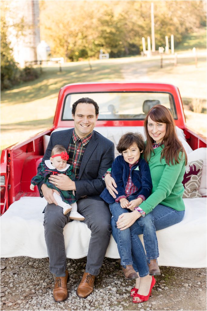 Family of 4 are seated in the back of a red truck. Dad is wearing a green and red plaid button up shirt and a black blazer and grey pants. Baby is wearing a green and red plaid dress with a white collar, white tights and shoes and a red bow. Son is wearing a red plaid shirt and a navy blue blazer and blue jeans with cowboy boots. Mom is in a long sleeve green sweater, blue jeans and red peep toe red heels.