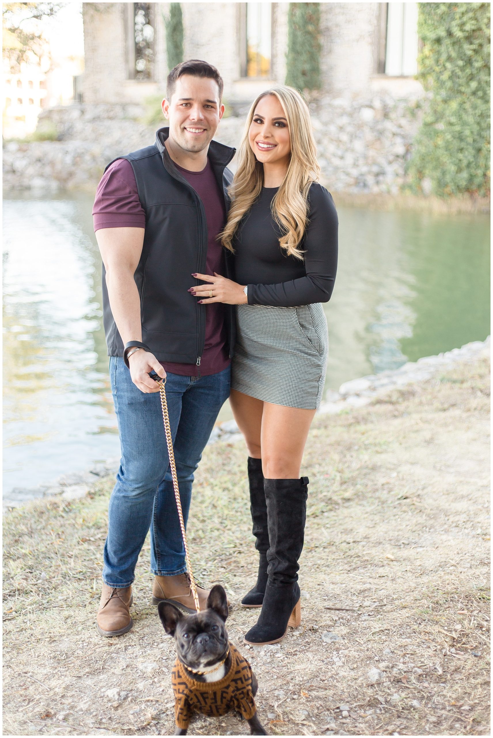 A gorgeous couple is just in front of a lake with their dog who is wearing a sweater. The husband is wearing a purple shirt with a grey vest and blue jeans. The wife is wearing a black long sleeve shirt and a black and white checkered skirt and black knee-high boots.