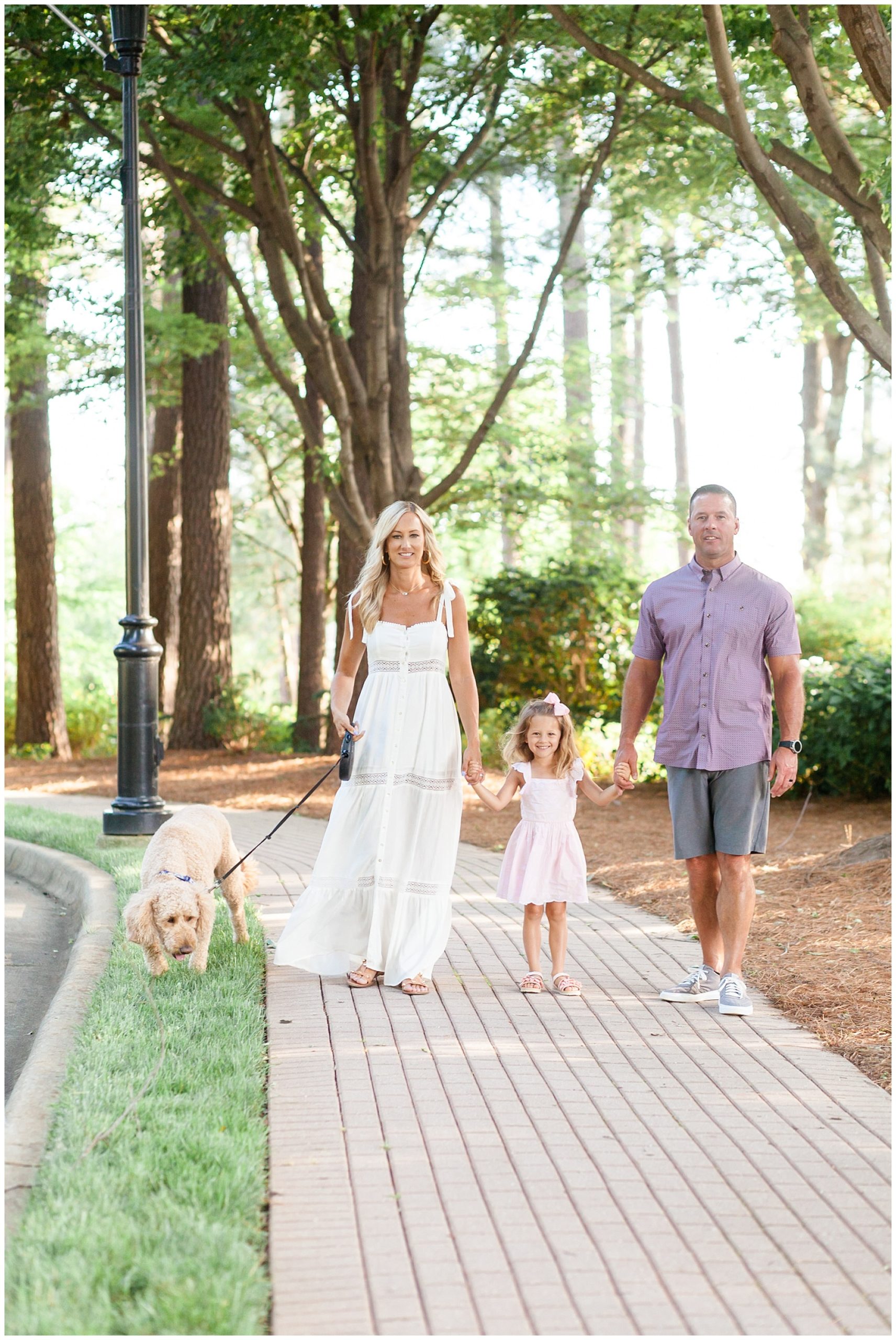 A beautiful family of 3 and their dog are walking down a brick walkway with trees just in the background. Mom is wearing a white ankle-length tiered sleeveless dress that ties at the shoulder. The daughter is wearing a white dress with flutter sleeves and a scalloped hem. Dad is wearing a purple short sleeve button-up shirt with grey shorts.