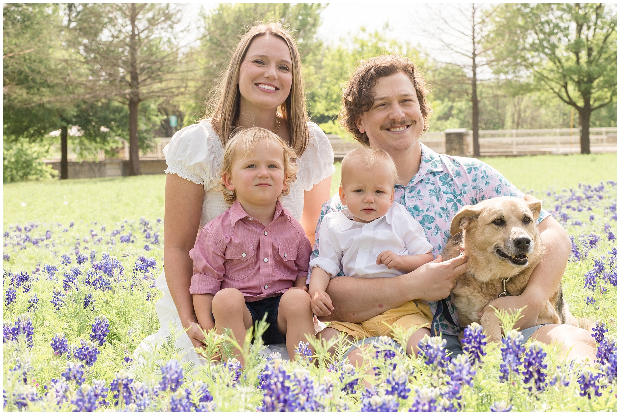 A beautiful family of 4 and their dog are sitting in a field of bluebonnets. Mom is wearing a short sleeve white shirt. 1 son is wearing a long sleeve button-up shirt rolled to the elbows and blue shorts. 2nd son is wearing a  long sleeve white shirt with sleeves rolled and mustard yellow pants. Dad is wearing a pink short sleeve shirt and green print.