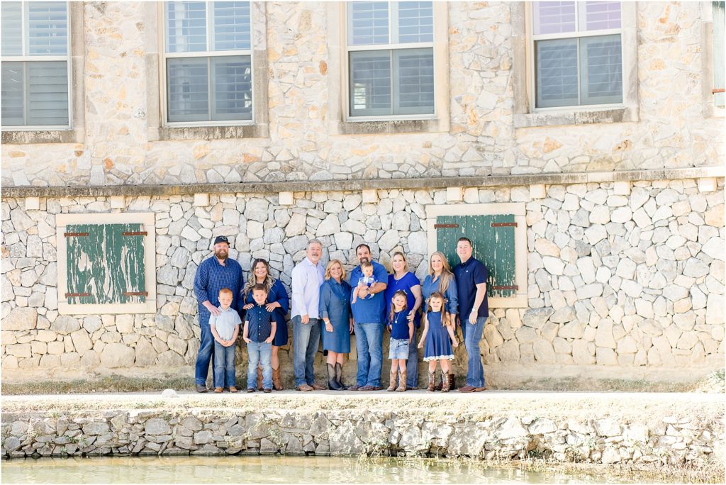 Family of 13 are standing in front of a rustic stone wall with green wooden shutters on the widows. The family is wearing several different shades of blue.